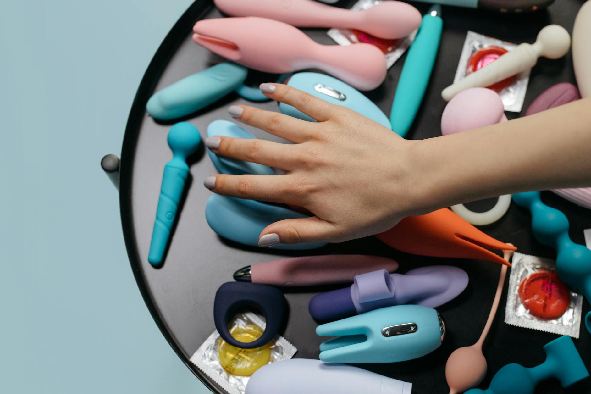 A hand reaching over a variety of colorful sex toys arranged on a table